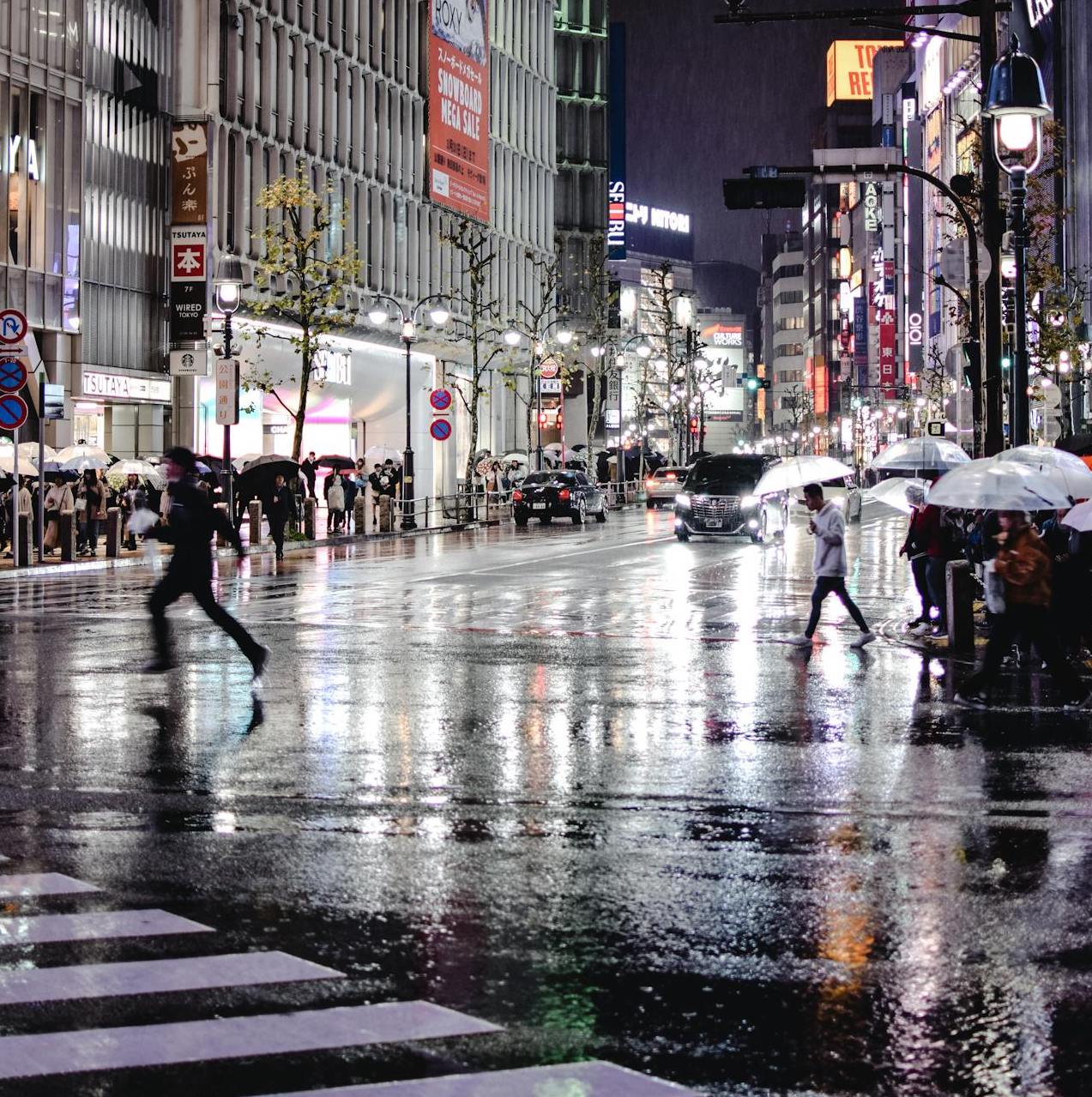 Shibuya Crossing at night, with people crossing the iconic intersection under umbrellas in the rain, surrounded by illuminated buildings and signs.